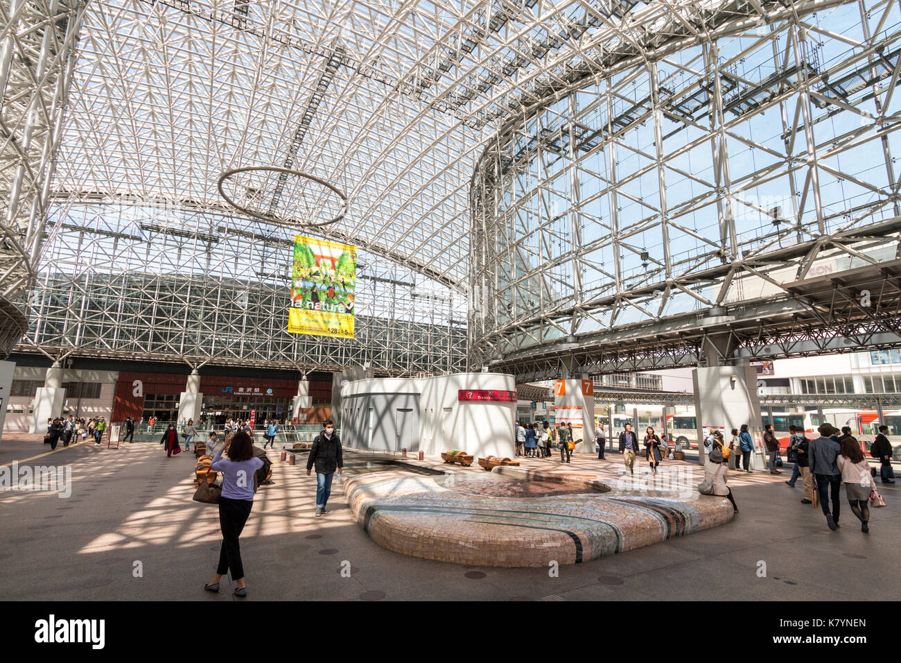 Kanazawa Station, Japan. Innenraum der berühmten Wahrzeichen Motenashi Kuppel mit großen Poster hängen, Leute, Sitzecke, tagsüber, blauer Himmel. Stockfoto
