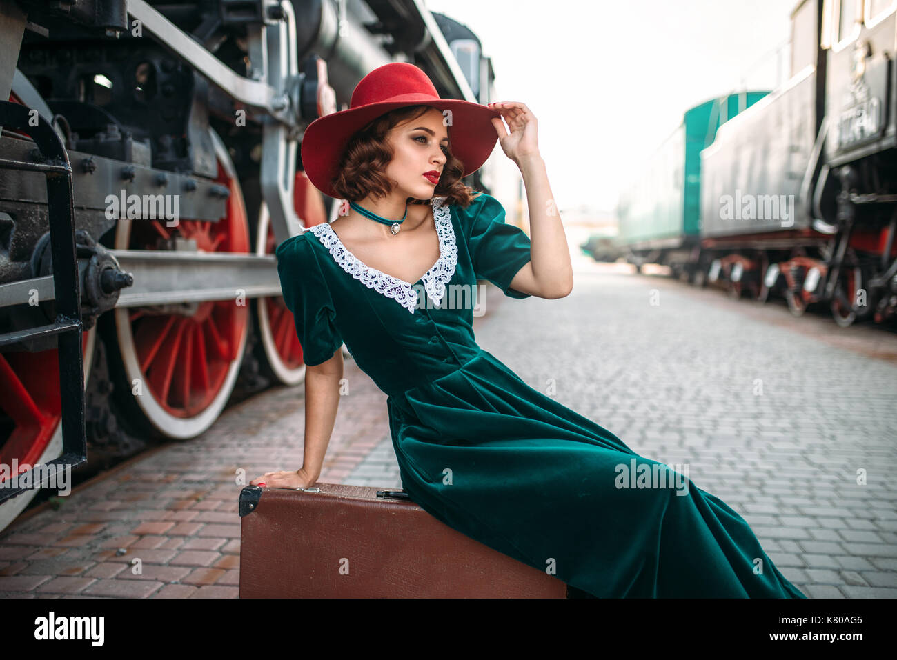 Junge altmodische Frau sitzt auf der Koffer gegen vintage Dampfzug, rote Räder Nahaufnahme. Alte Lokomotive. Lokomotive, Reisen per Bahn Stockfoto