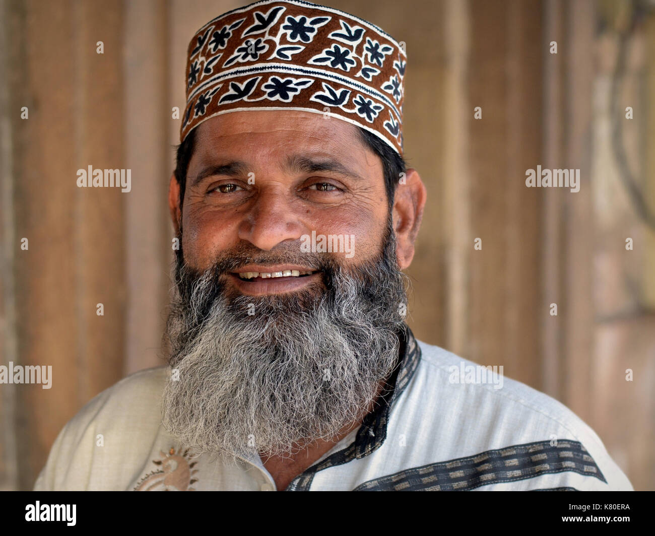 Der indische muslimische Mann mit einem im muslimischen Stil gestutzten Vollbart trägt eine gemusterte islamische Betkappe (taqiyah) im omanischen Stil und lächelt für die Kamera. Stockfoto