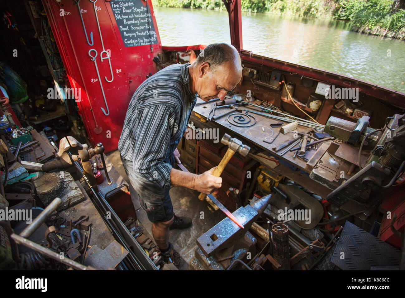 Hohe Betrachtungswinkel der Schmied an seiner Werkbank mit seinem Boot auf dem Wasser, Hämmern von heißem Metall. Stockfoto