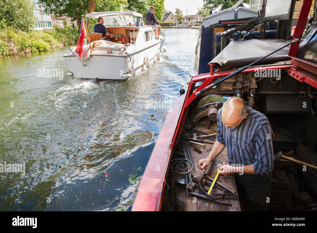 Hohe Betrachtungswinkel von einem Schmied bei seiner Bank arbeitet an einem NARROWBOAT auf dem Wasser, mit einem Maßband. Ein Motorboot im Gange. Stockfoto