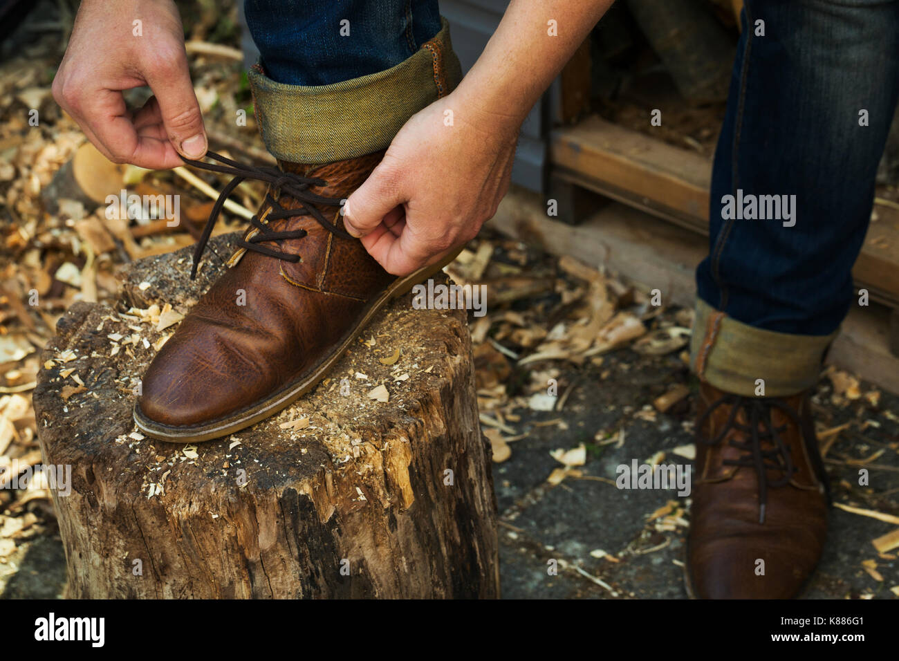 Ein Mann in Jeans mit turnups sitzend beugte seine Schnürsenkel seiner braunen Leder Schuhe zu binden, einen Fuß auf dem Verteilerblock. Stockfoto