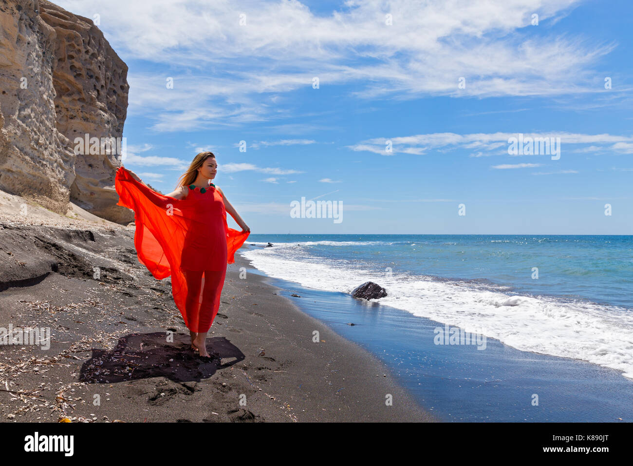 Junge Frau im roten Kleid und geht barfuß auf dem schwarzen Sand Meer Stockfoto