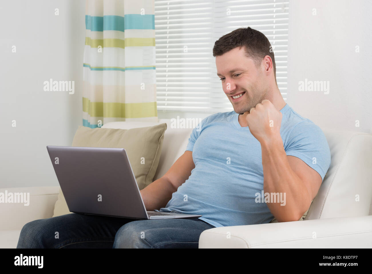Lächelnd Mitte der erwachsene Mann ballte die Faust, während mit Laptop auf dem Sofa zu Hause Stockfoto