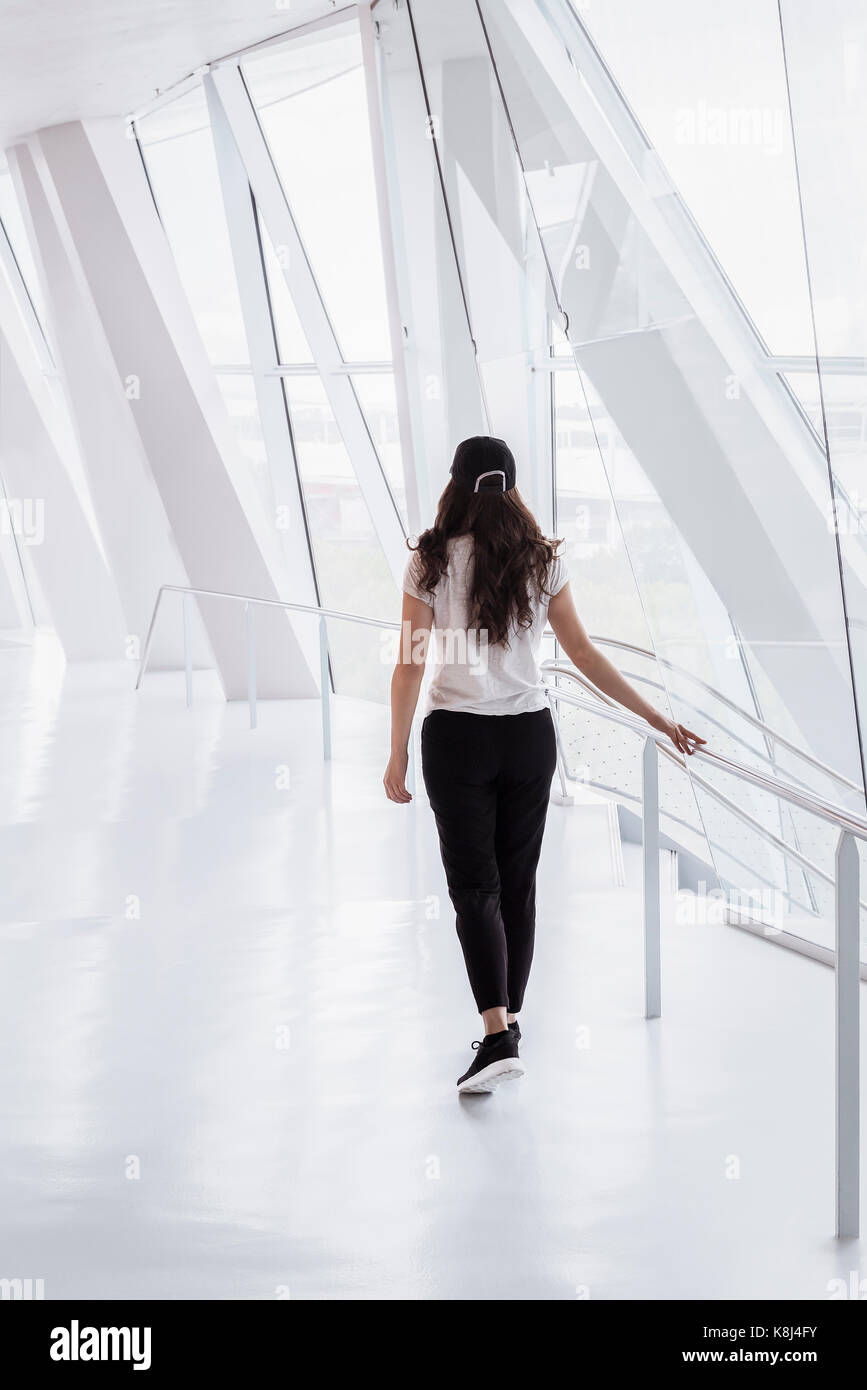 Junge Frau, lässig gekleidet, Spaziergänge auf einem hellen, weißen Flur, von einem Bürogebäude mit hohen Fenstern und modernen Spalten. Stockfoto