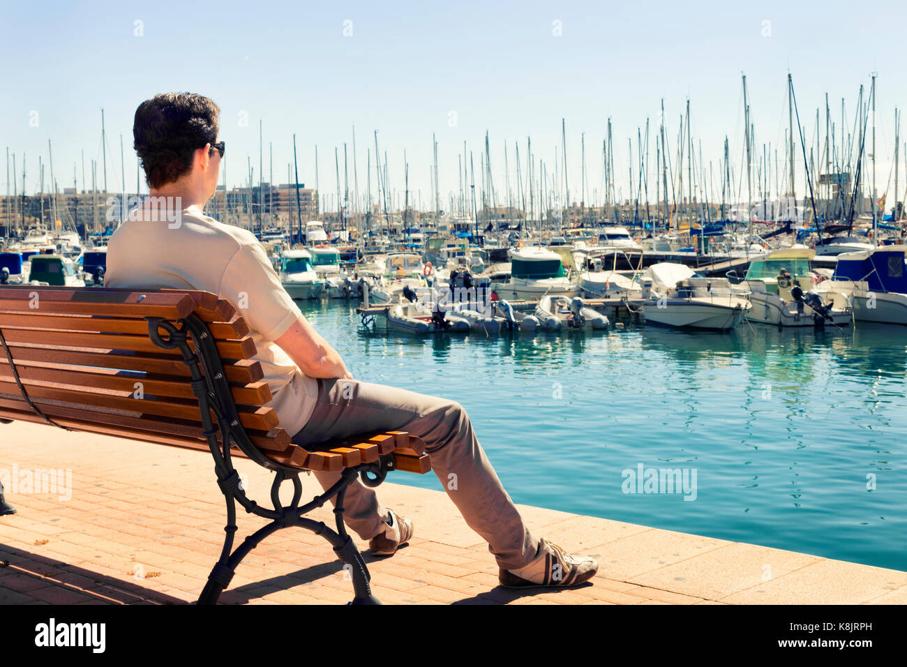 Mann sitzt und Sonnenbaden auf einer Bank der Betrachtung der Boote im Hafen von Alicante. Spanien Stockfoto