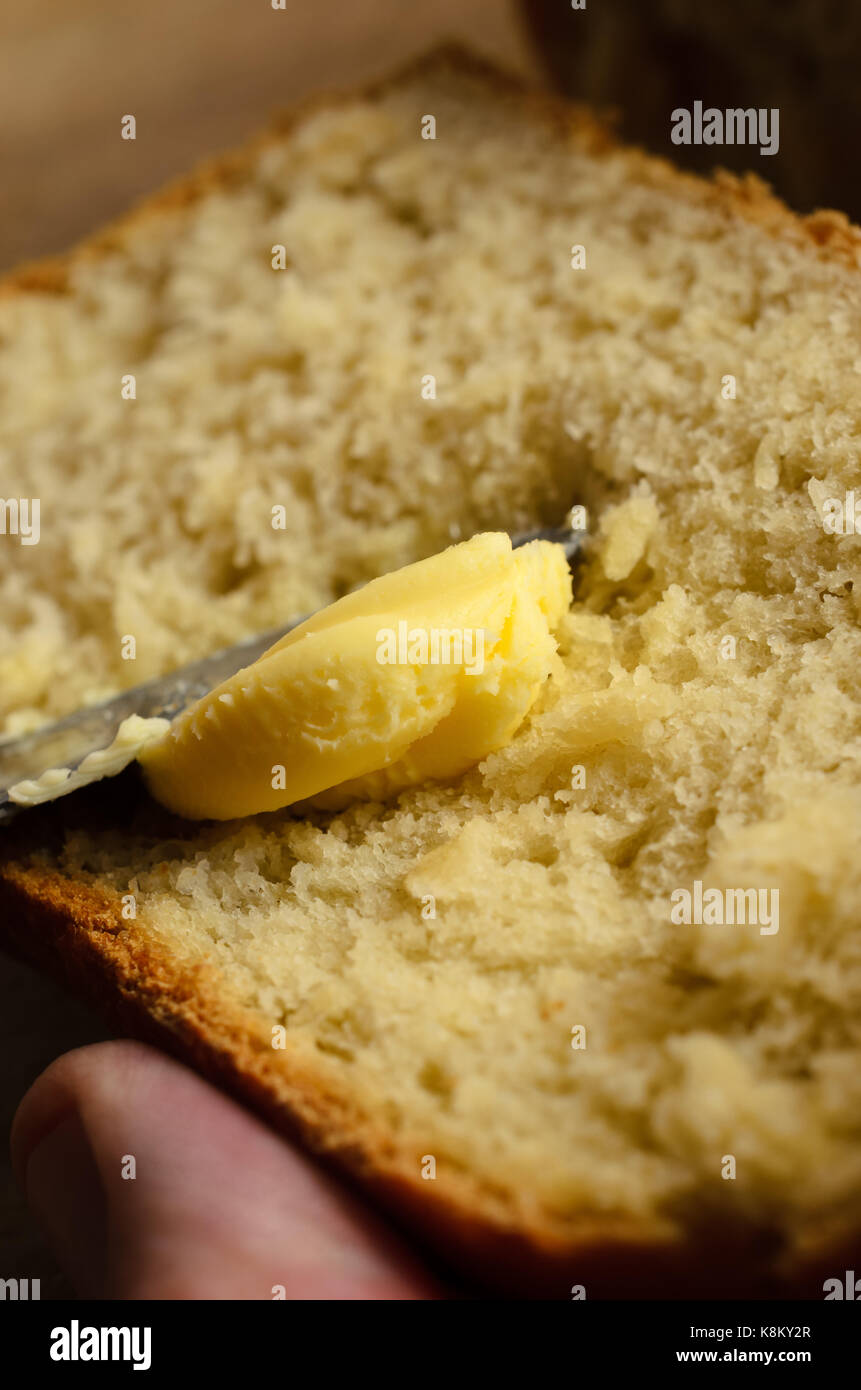 Nahaufnahme von echten Butter verteilen sich auf eine Scheibe frisch gebackenes Brot, von männlichen Hand mit Daumen im Rahmen statt. Brot ist nur im Hintergrund sichtbar. Stockfoto