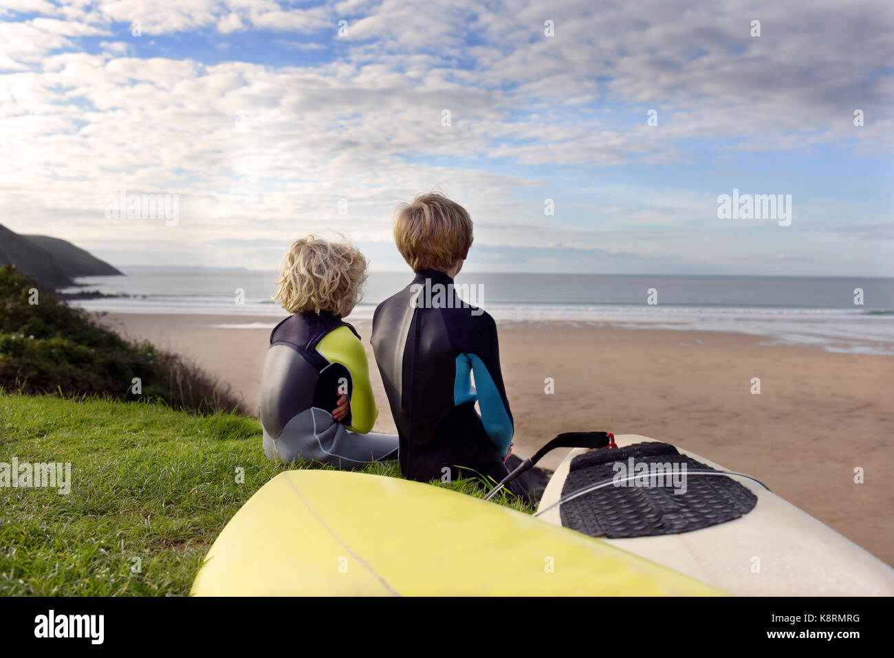 Zwei junge Brüder Blick zum Meer wie Betrachten Sie sitzen auf Gras Hang mit Blick auf den Strand tragen Neoprenanzug neben ihren eigenen Junior surfbo Stockfoto