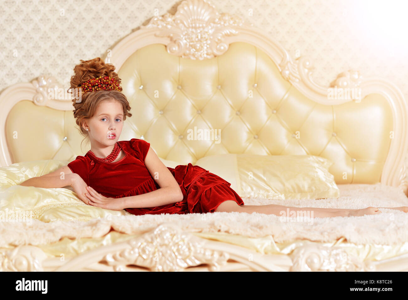 Süße kleine Mädchen in roten Samt Kleid mit retro Frisur liegen auf beige Couch Stockfoto