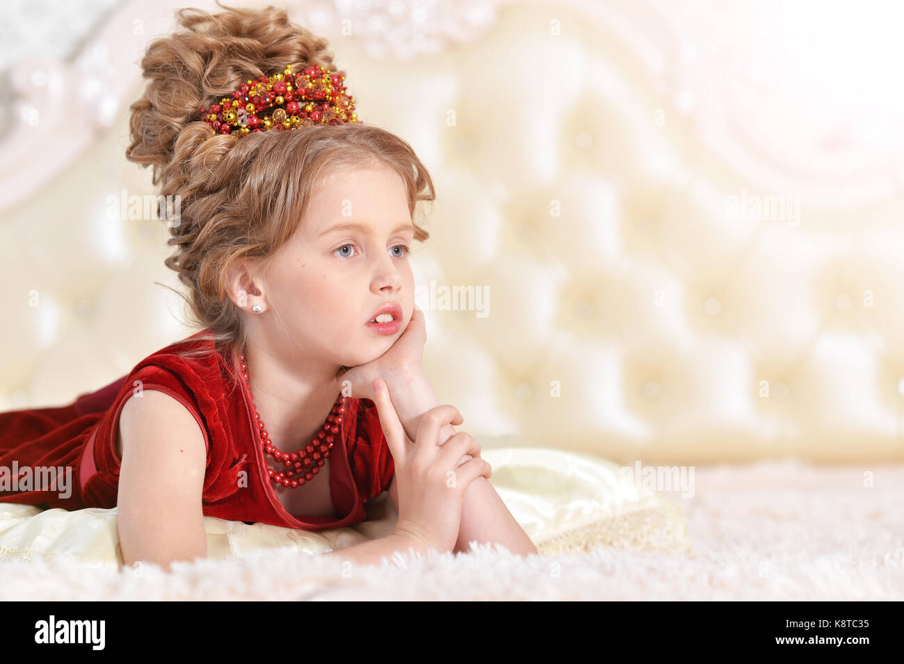 Süße kleine Mädchen in roten Samt Kleid mit retro Frisur liegen auf beige Couch Stockfoto