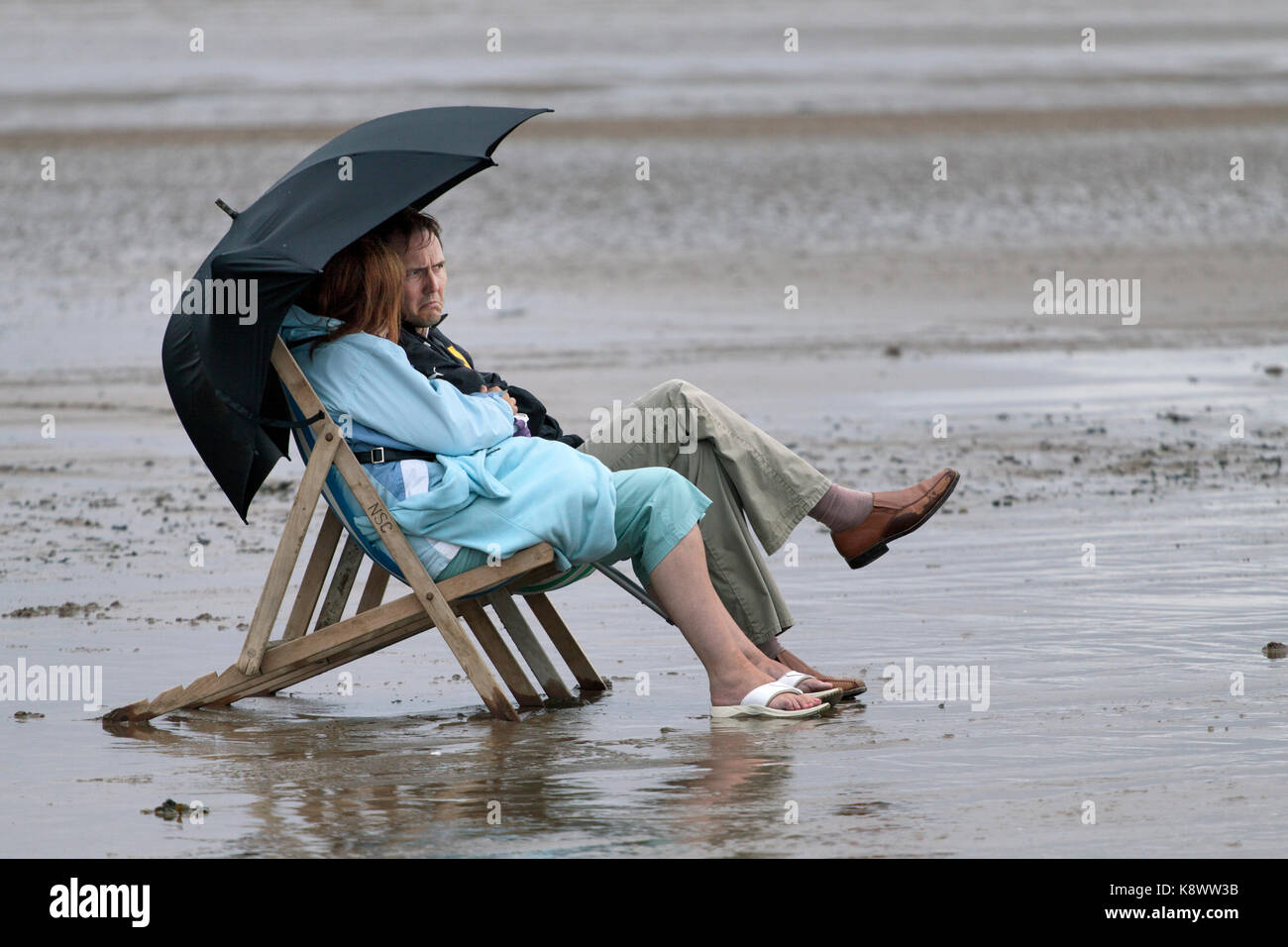 Ein Paar mittleren Alters saß in Liegestühlen am Strand bei einem regen Sturm. Beide tragen Kleider und die Stühle mit einem Regenschirm abgedeckt Stockfoto