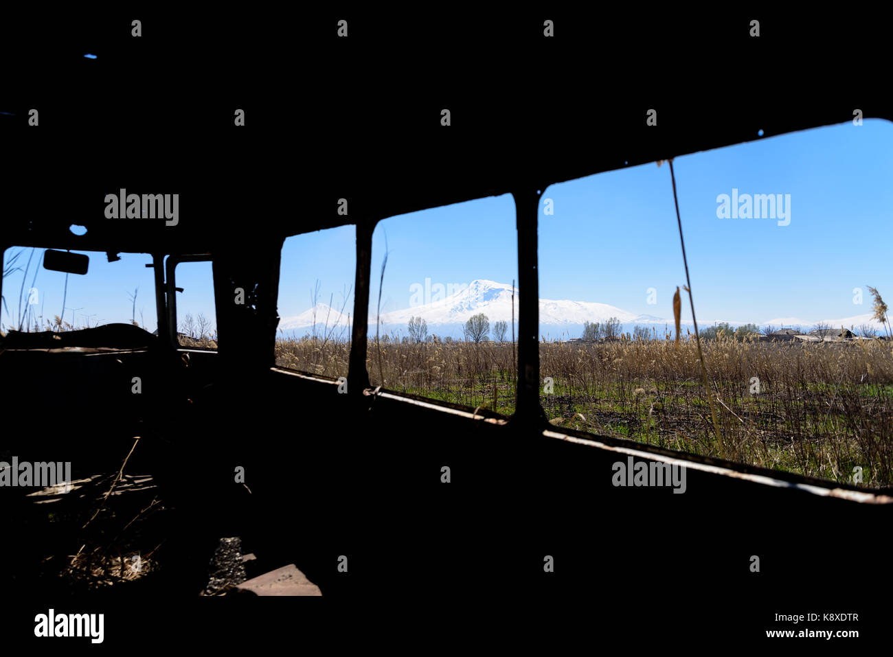 Blick von innen auf einen verlassenen und rostigen alten Sowjet Russischer Bus in der Mitte von Schilf und Landwirtschaft Felder Mit schneebedeckter landschaftlicher Ararat-Berg Stockfoto