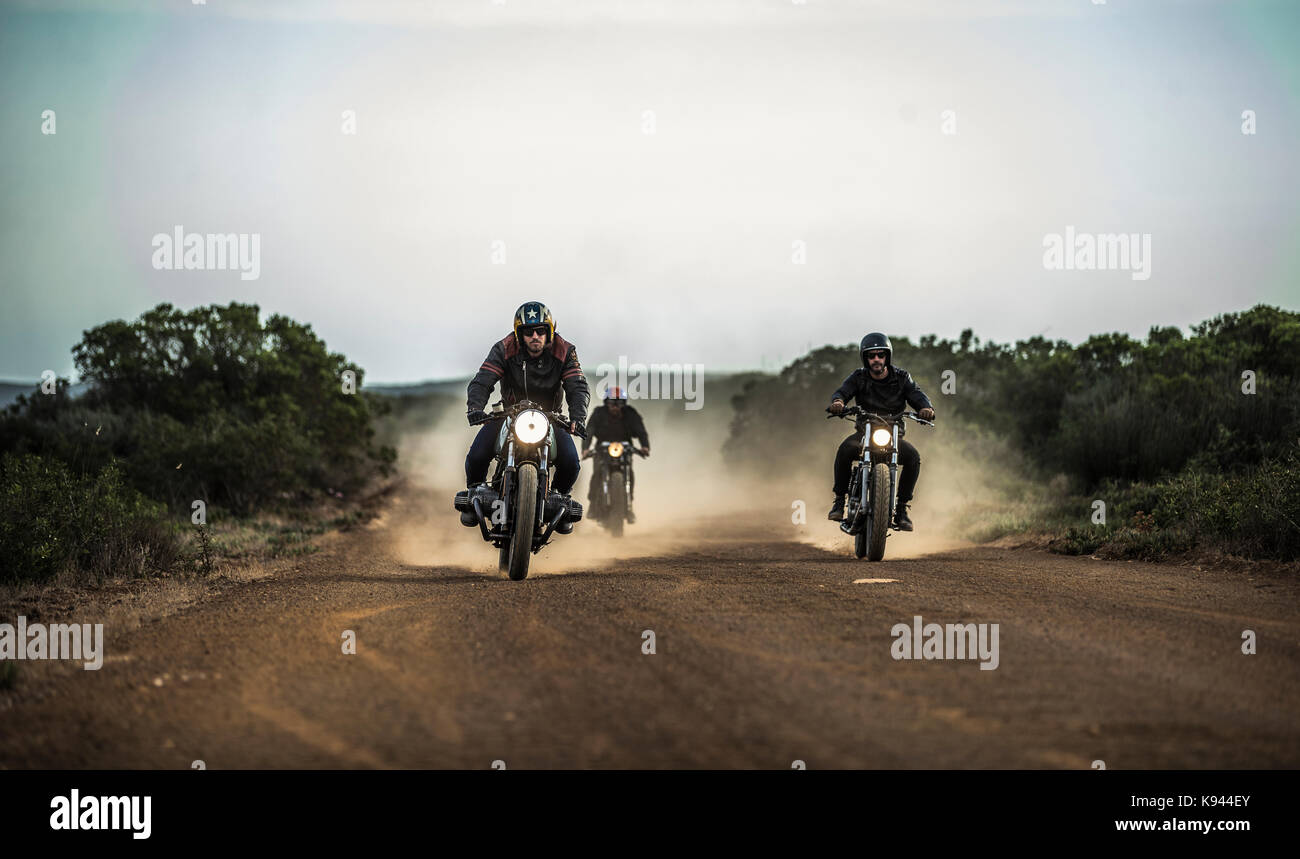 Drei Männer reiten Café Racer Motorrad entlang der staubigen Piste. Stockfoto