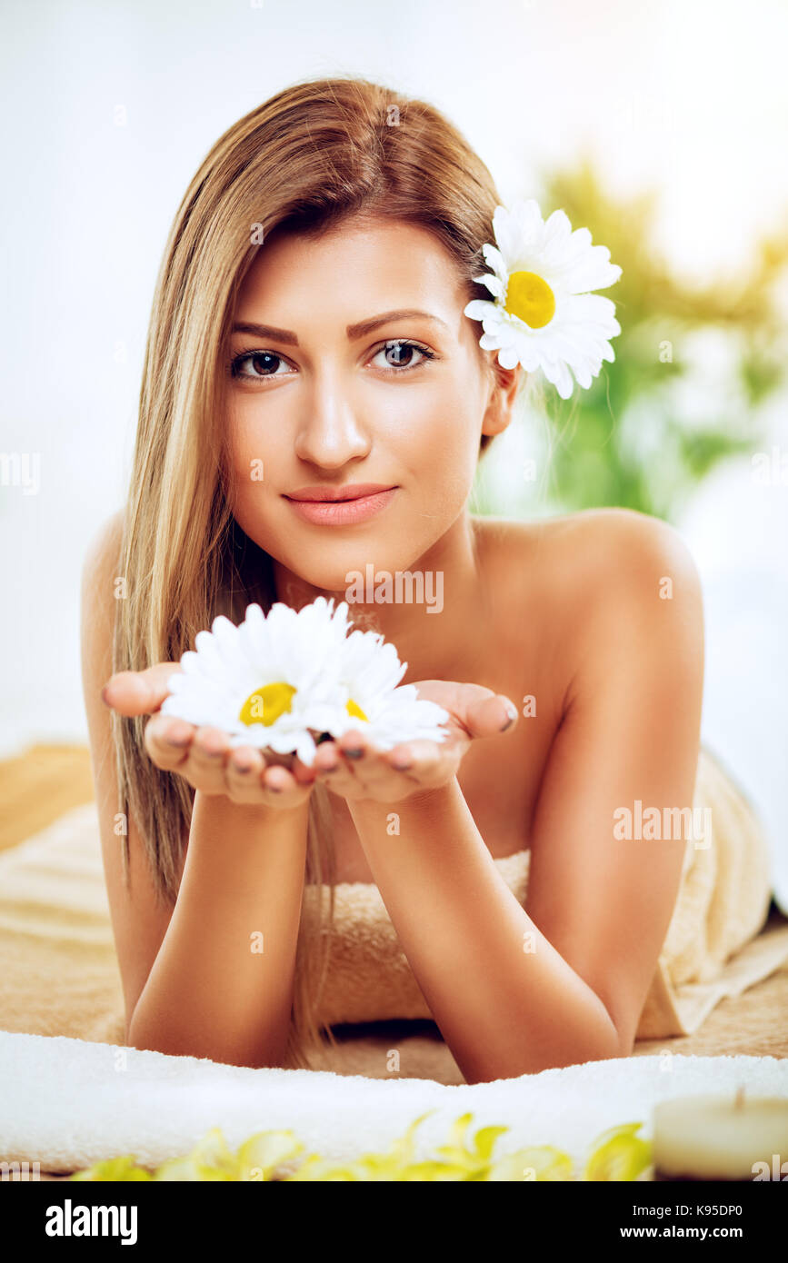 Nette junge lächelnde Frau genießen Sie während einer skincare Behandlung in einem Spa. Sie schaut an Kamera und halten weiße Blumen in den Händen. Stockfoto