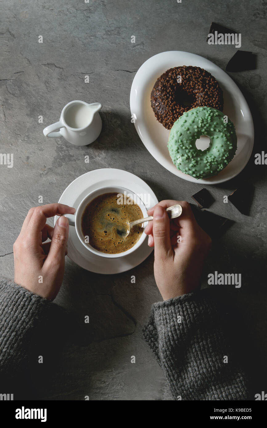 Platte von Bunt glasierter Krapfen mit gehackte Schokolade, Tasse schwarzen Kaffee, Kanne Milch über grau Textur Tabelle. Weibliche Hände. Flach mit dem Raum. R Stockfoto