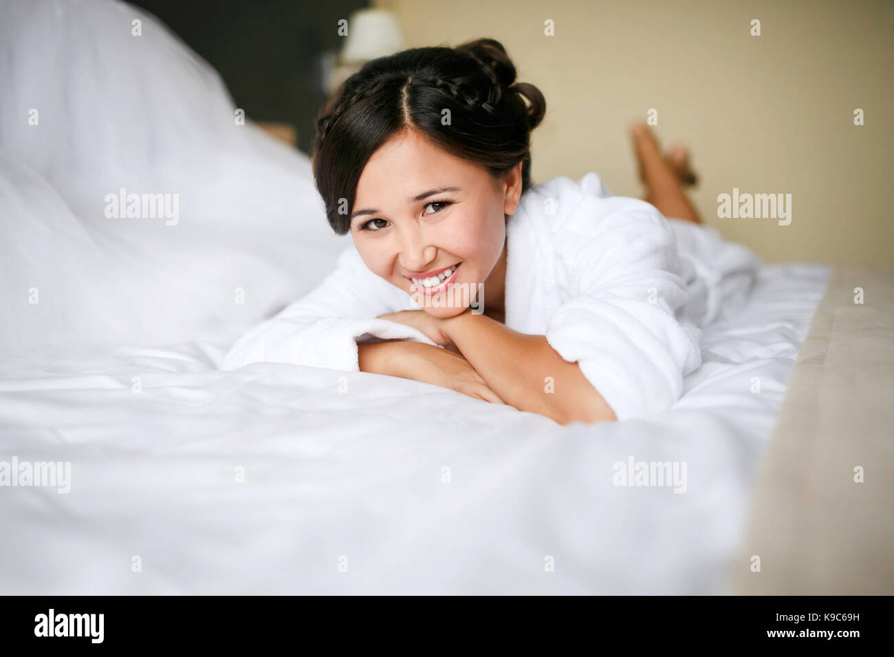 Frohe Braut morgens im Bett im Bademantel lächelt Fröhlich, positiv, mit einem fertigen Frisur und Make-up, ein Brautkleid im Hintergrund. In Erwartung der Ehe. Stockfoto