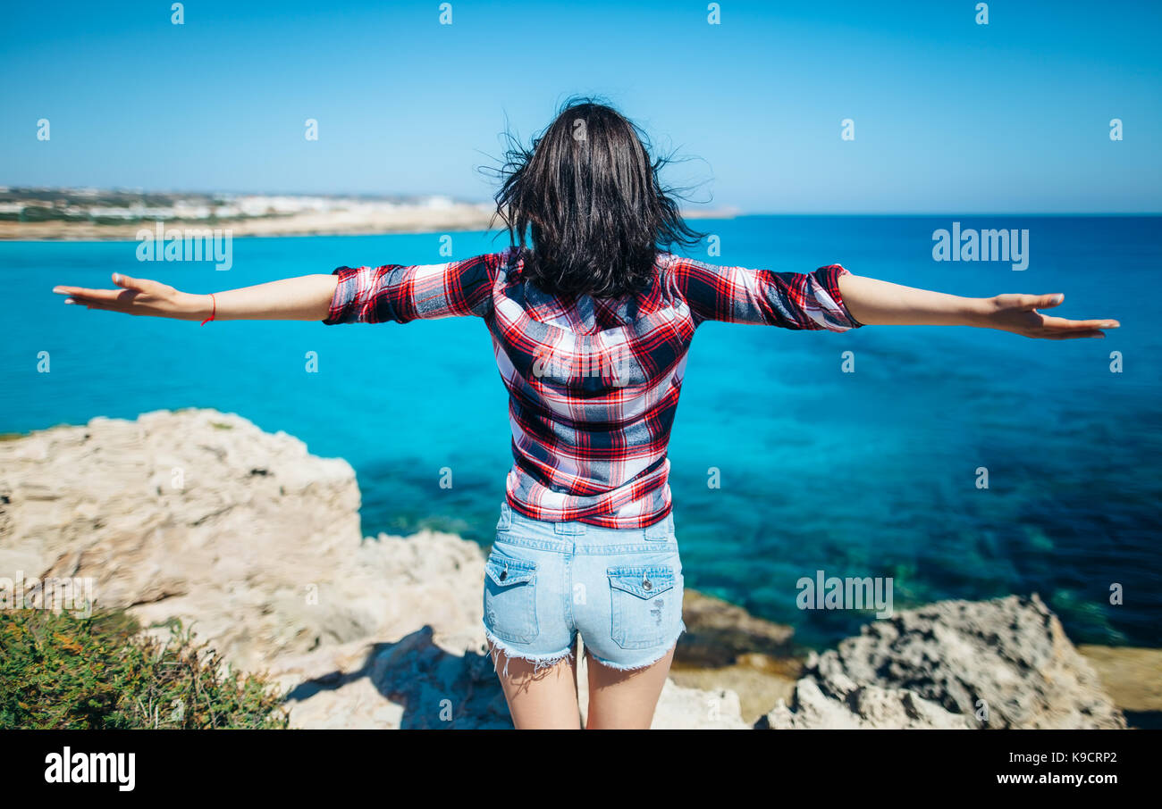 Rückansicht der jungen Frau mit dem Wind im Haar ihre Hände heben auf der Klippe über dem Meer. Entspannen Konzept. Stockfoto