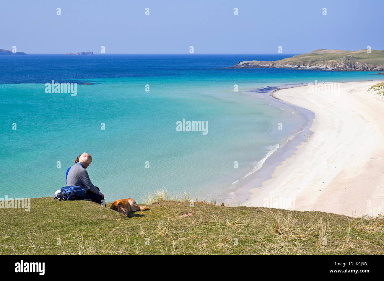 Zwei Damen mittleren Alters mit Hund in Picknick am Rande der grasigen Felsen mit Blick auf die ruhige See und leeren weißen Sandstrand, balnakeil Bay, Sutherland, Großbritannien Stockfoto