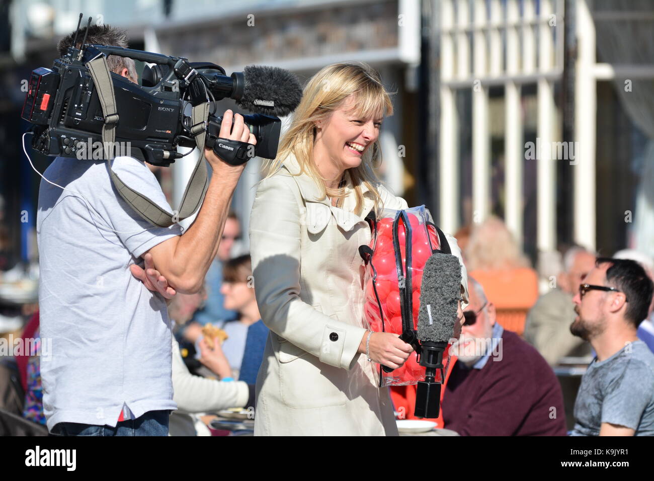 Outside Broadcast an einem heißen Tag zeigen einen hübschen weiblichen Reporter lachend während der Durchführung eines Interviews, durch ein TV-Kameramann gefilmt wird, in Großbritannien. Stockfoto