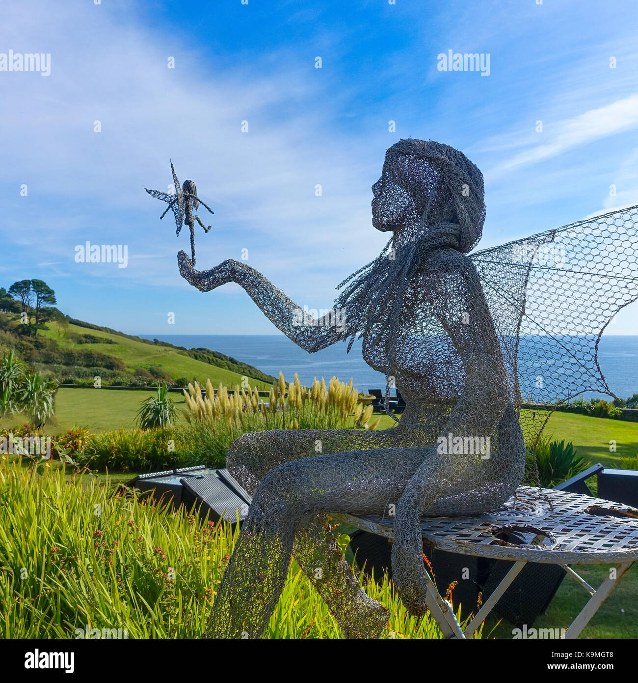 Lebensgroße Drahtskulptur eines Engels oder einer Feenkönigin mit Flügeln, die auf einem Gartentisch sitzen und eine winzige Fee in der Hand halten, die ihren ersten Flug macht. Stockfoto Lebensgroße Drahtskulptur eines Engels oder einer Feenkönigin mit Flügeln, die auf einem Gartentisch sitzen und eine winzige Fee in der Hand halten, die ihren ersten Flug macht. Stockfoto