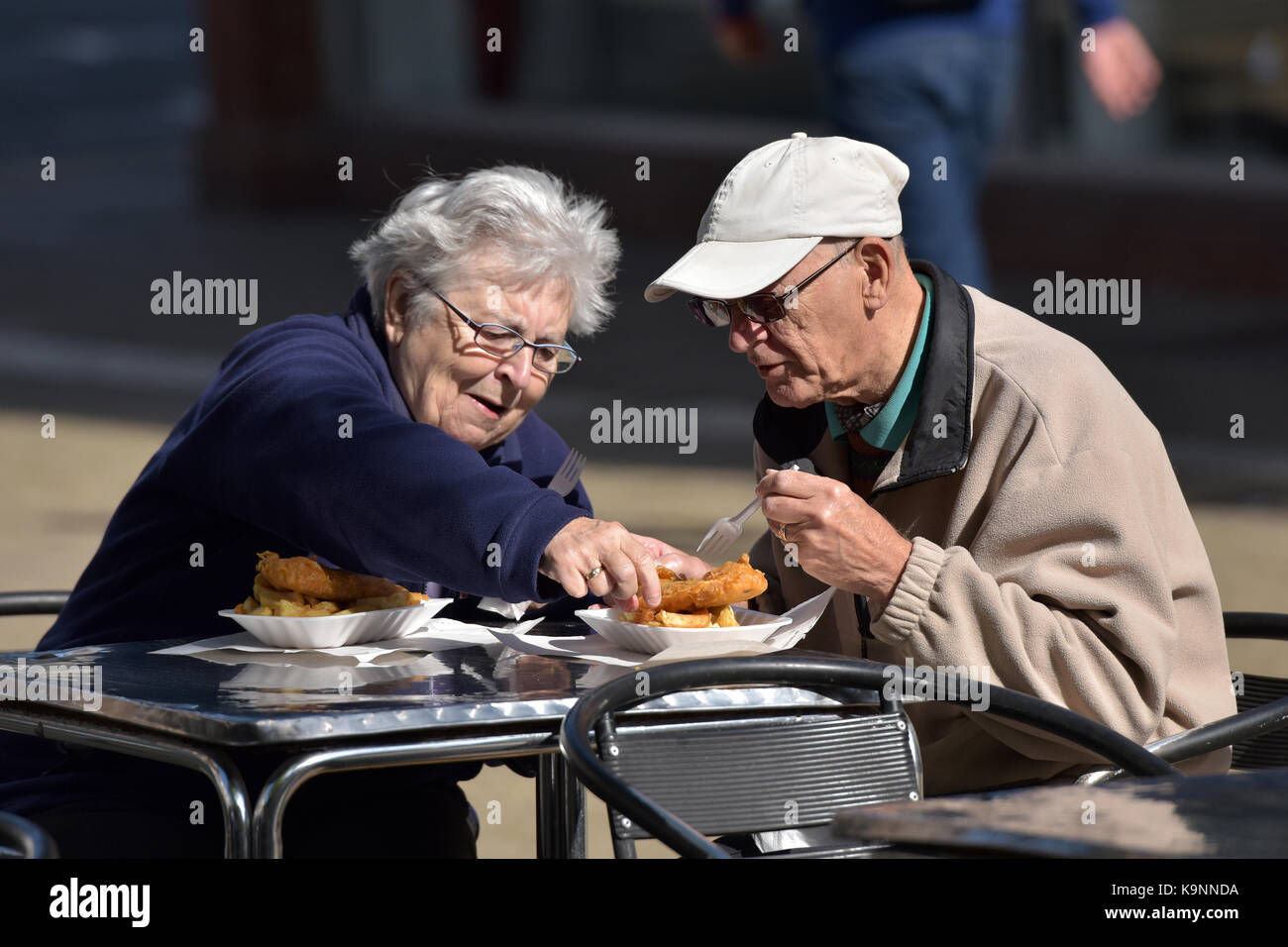 Ein älterer Mann und Frau oder Mann und Frau Fisch und Chips auf einen Tisch draußen Essen im Freien mit Lady stehlen oder Kneifen ein Chip vom Ehemann. Stockfoto