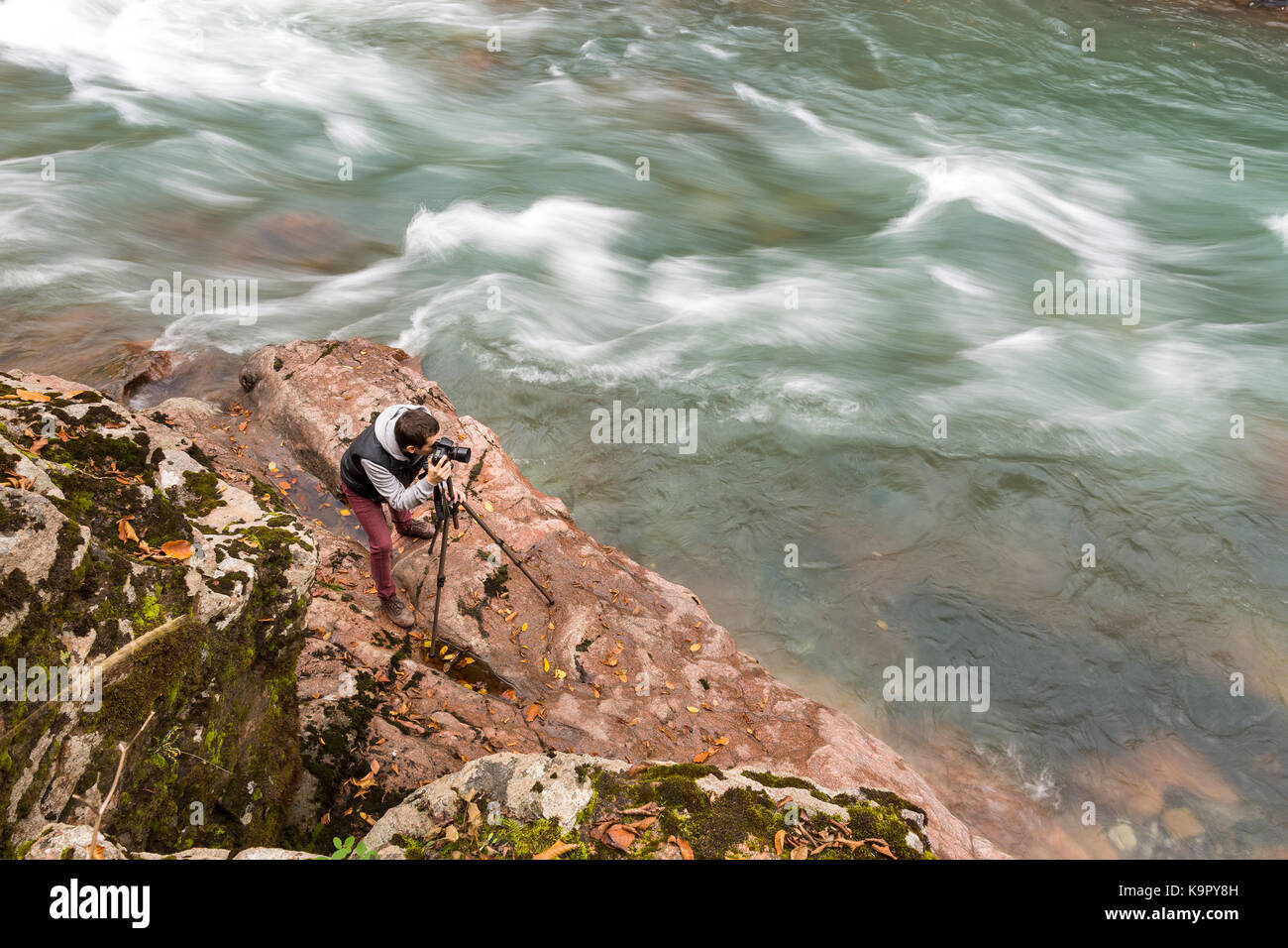 Hobby, hohe Qualifikation, Natur Konzept. professioneller Fotograf am Werk ist, stehend mit Stativ an der felsigen Küste in der Nähe von Fluss mit Kabbelwasser, fließt sehr schnell Stockfoto