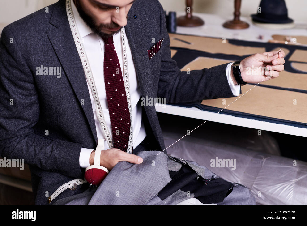 Junge Schneider in männlichen Jacke Nähen mit Nadel und Faden beim Sitzen am Tisch gewickelt, Interieur des Atelier auf Hintergrund Bärtigen Stockfoto