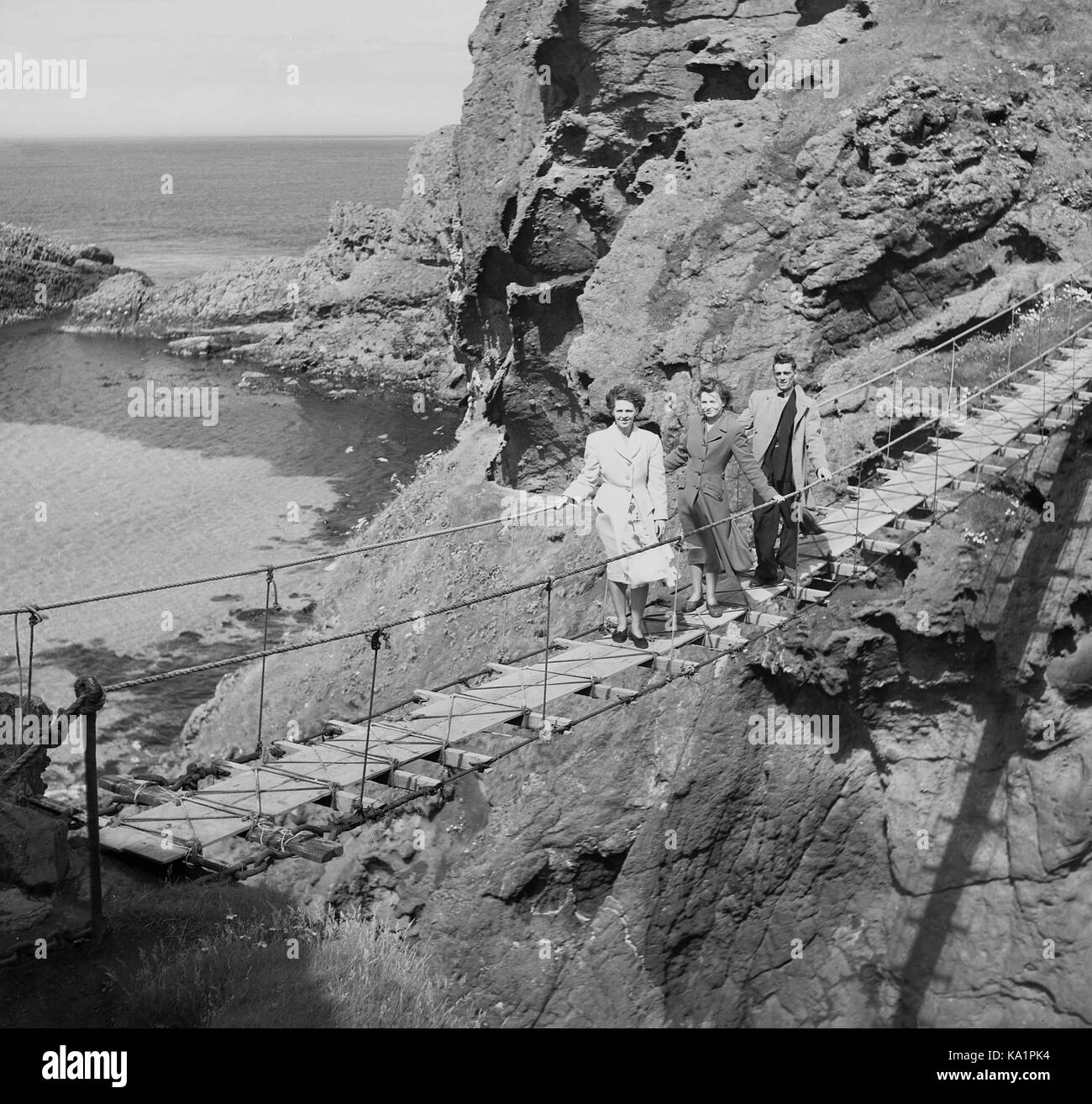 Ein Mann und zwei Damen aus den 1950er Jahren, die auf der Carrick-a-Rede-Seilbrücke, einer berühmten Seil- oder Schaukelbrücke in der Nähe von Ballintoy in der Grafschaft Antrim, Nordirland, stehen, die erstmals 1755 vom lokalen Lachsfischer errichtet wurde. Stockfoto