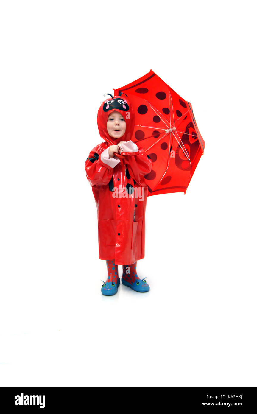 Das liebenswerte kleine Mädchen trägt einen roten, pfannenförmigen Regenmantel und Regenschirm. Sie macht ein lustiges Gesicht und steht in einem ganz weißen Raum. Stockfoto