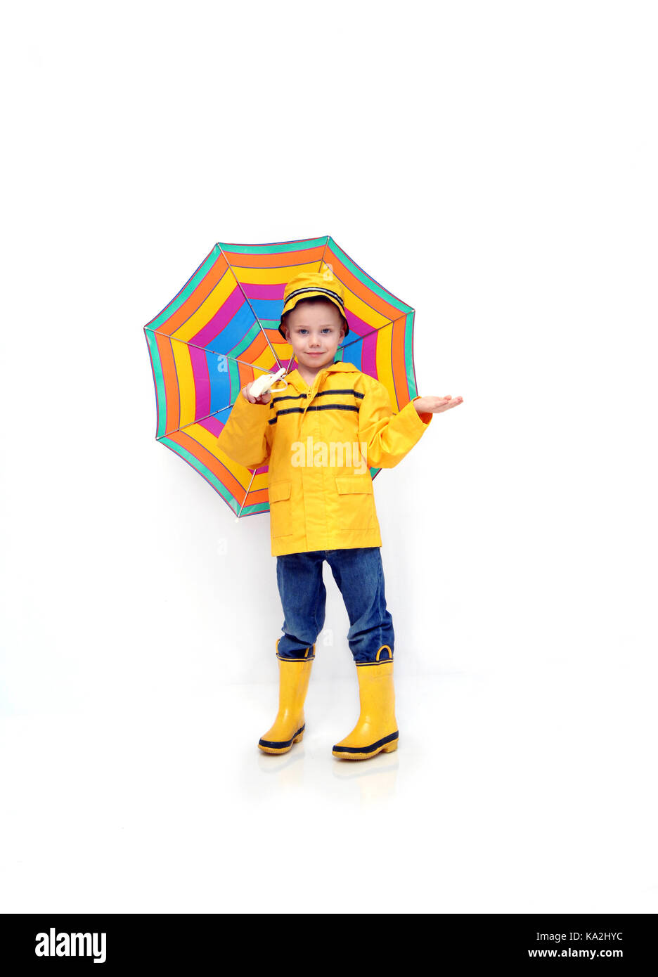 Little boy Kontrollen für Regen, während in einem gelben Slicker und Gummistiefel und Hut bekleidet. Er ist ein multi-gestreifte Schirm in einem weißen Raum. Stockfoto
