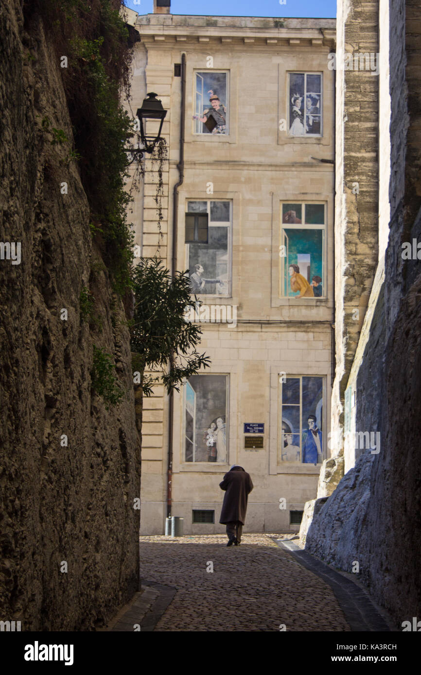 Eine einzelne männliche Figur Spaziergänge in einer schattigen Straße in Richtung eines Gebäudes mit Trompe-l'oeil-windows vergangener Zeiten. Stockfoto