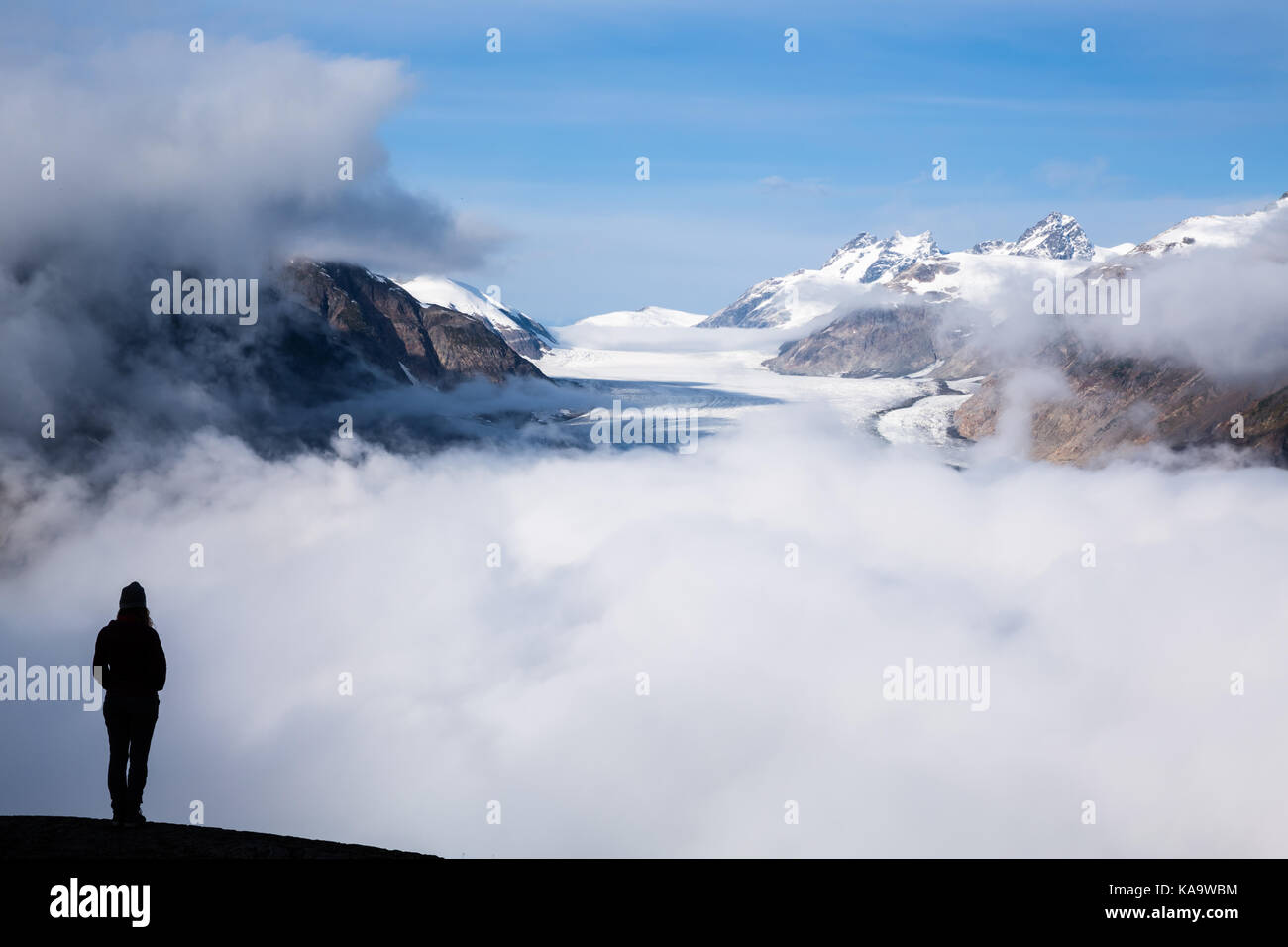 Kurz nach Sonnenaufgang, ein beschlagener Salmon Gletscher Gipfel hinter den Wolken. Die Sonne noch nicht erleuchteten Die übersehen, wo eine junge Frau steht looki Stockfoto