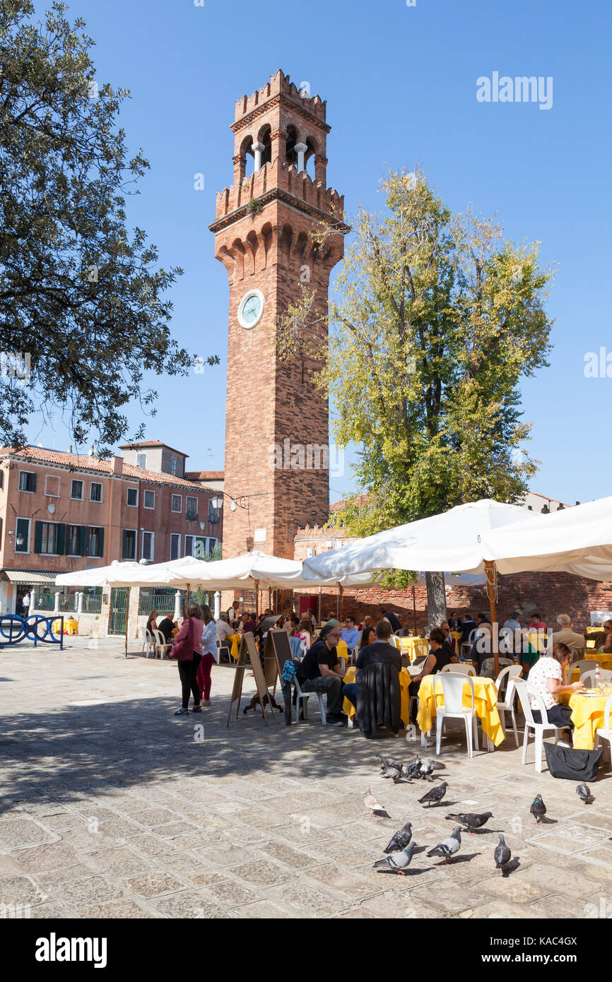 Touristen in Campo Santa Stefano, Murano, Venedig, Italien, Essen unter freiem Himmel Regenschirme mit dem 19. Jahrhundert Clock Tower hinter und Tauben, die in der f Stockfoto