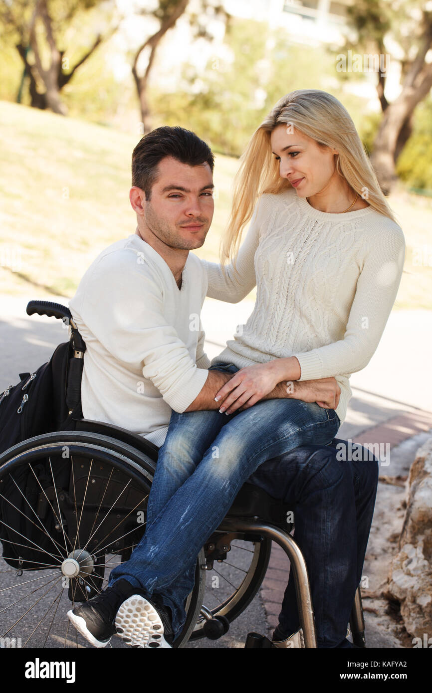 Junges Mädchen sitzt auf dem Schoß von ihrem behinderten Freund in einem Rollstuhl im Park Stockfoto