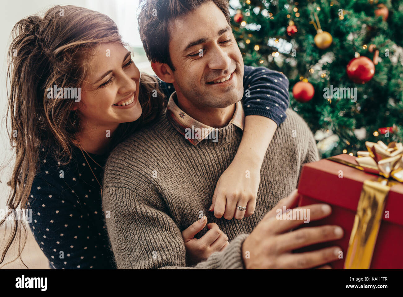 Paar mit Geschenkbox sitzen in der Nähe der Weihnachtsbaum. Lächelnde Frau ihrem Partner Holding von hinten, während die Ausgaben glückliche Momente Weihnachten feiern. Stockfoto