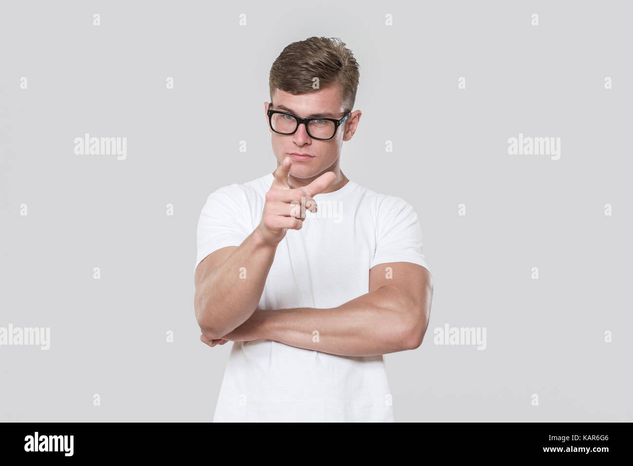 Hübscher junger Mann an der Kamera zeigt. Bild in einem Studio mit grauem Hintergrund genommen. Das Modell eine Brille trägt und ein weißes T-Shirt. Stockfoto