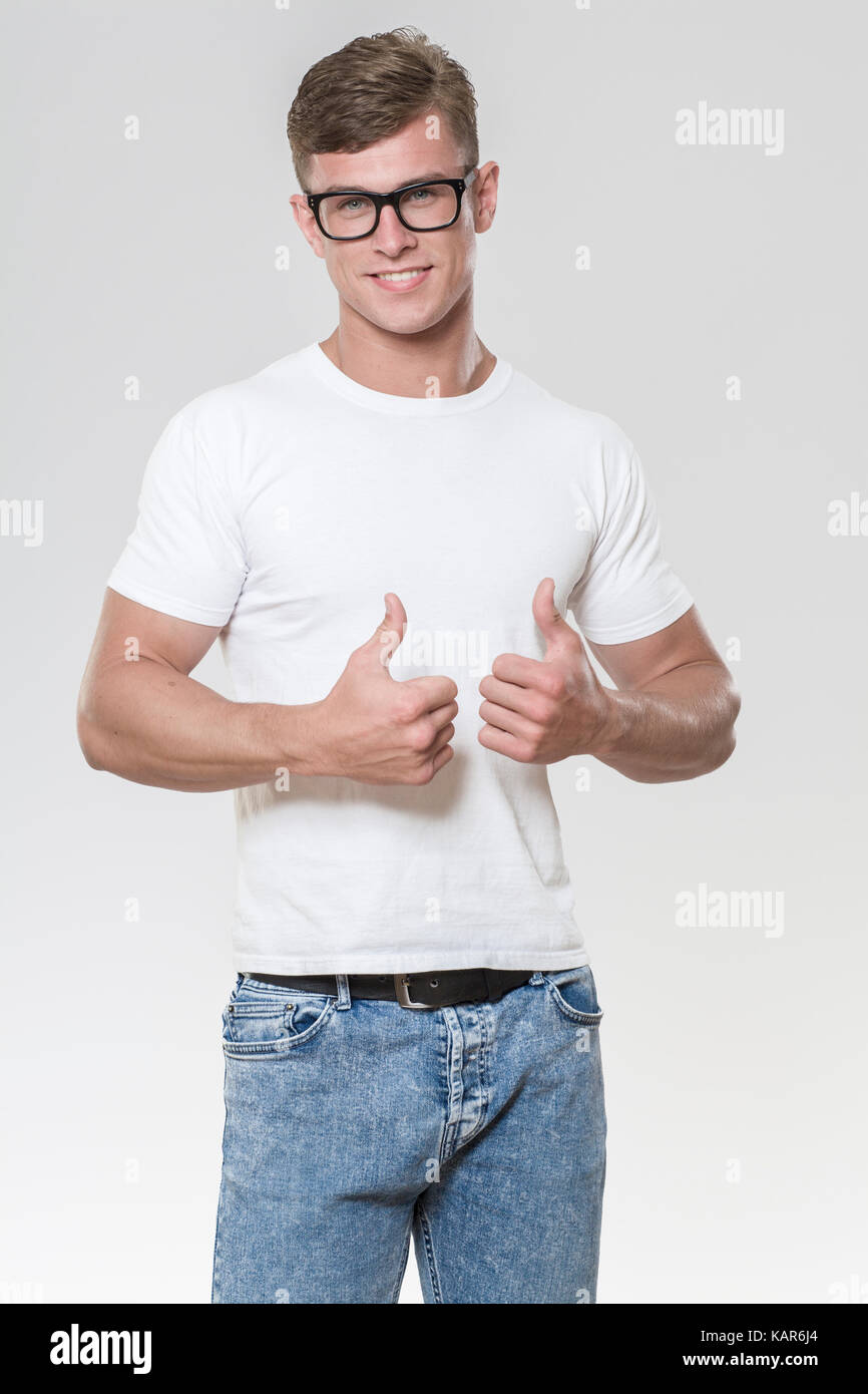 Lächelnd hübscher junger Mann mit Daumen nach oben. Bild in einem Studio mit grauem Hintergrund genommen. Das Modell eine Brille trägt und ein weißes T-Shirt. Stockfoto