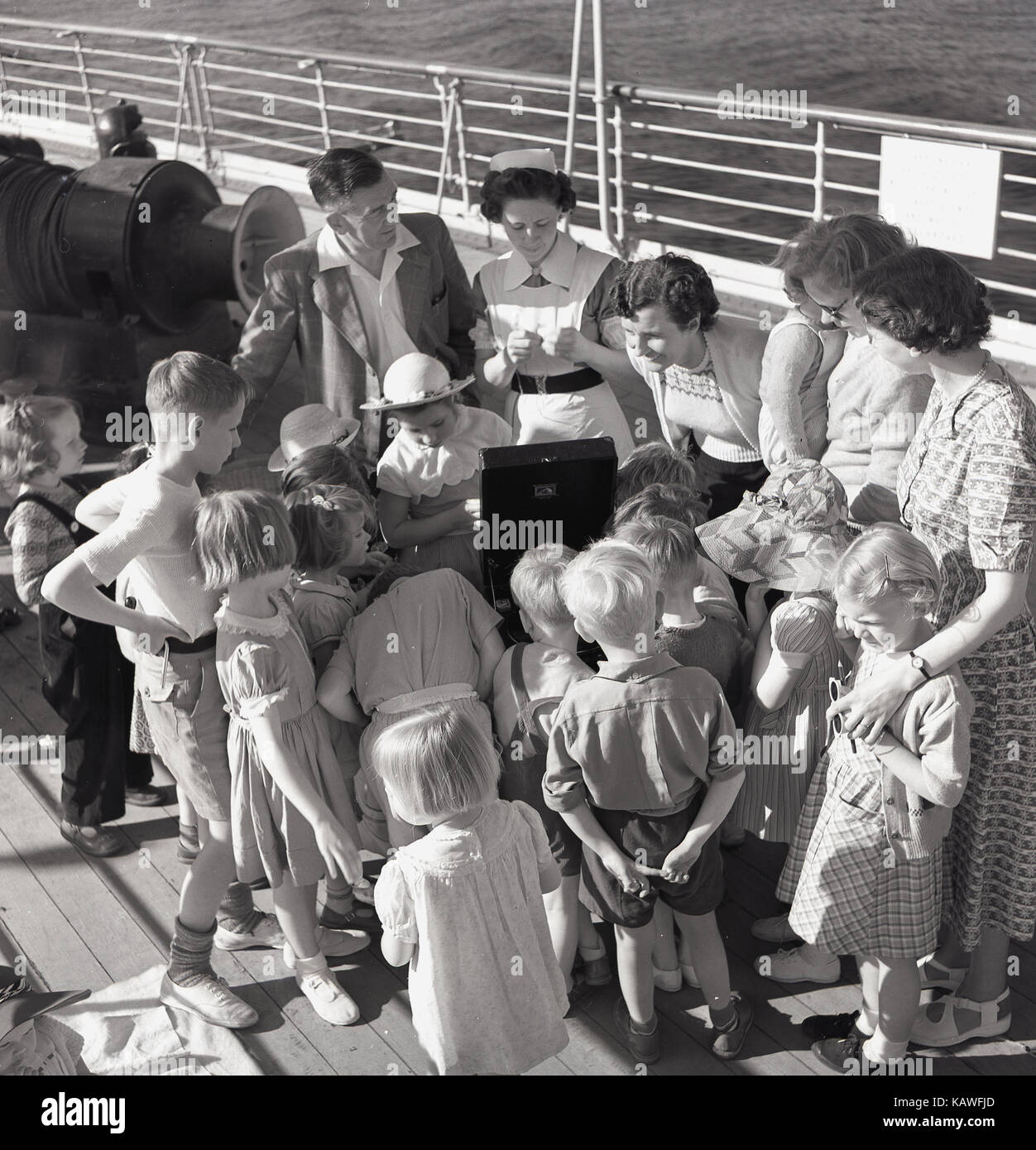1950, historische, junge Kinder Segeln auf einem Dampfschiff Union-Castle sammeln auf dem Deck um ein grammophon oder Plattenspieler mit einem stationsschwester und Eltern anwesend, um Musik zu hören. Stockfoto