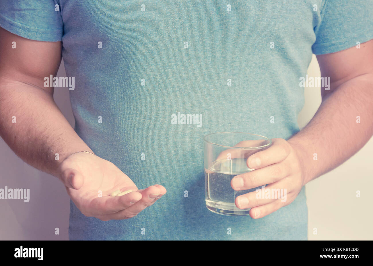 Ein junger Mann im blauen T-Shorts hält eine Gruppe von Kapseln mit Medizin oder Vitamine in seinen Händen und einem Glas Wasser in der anderen Hand. Stockfoto