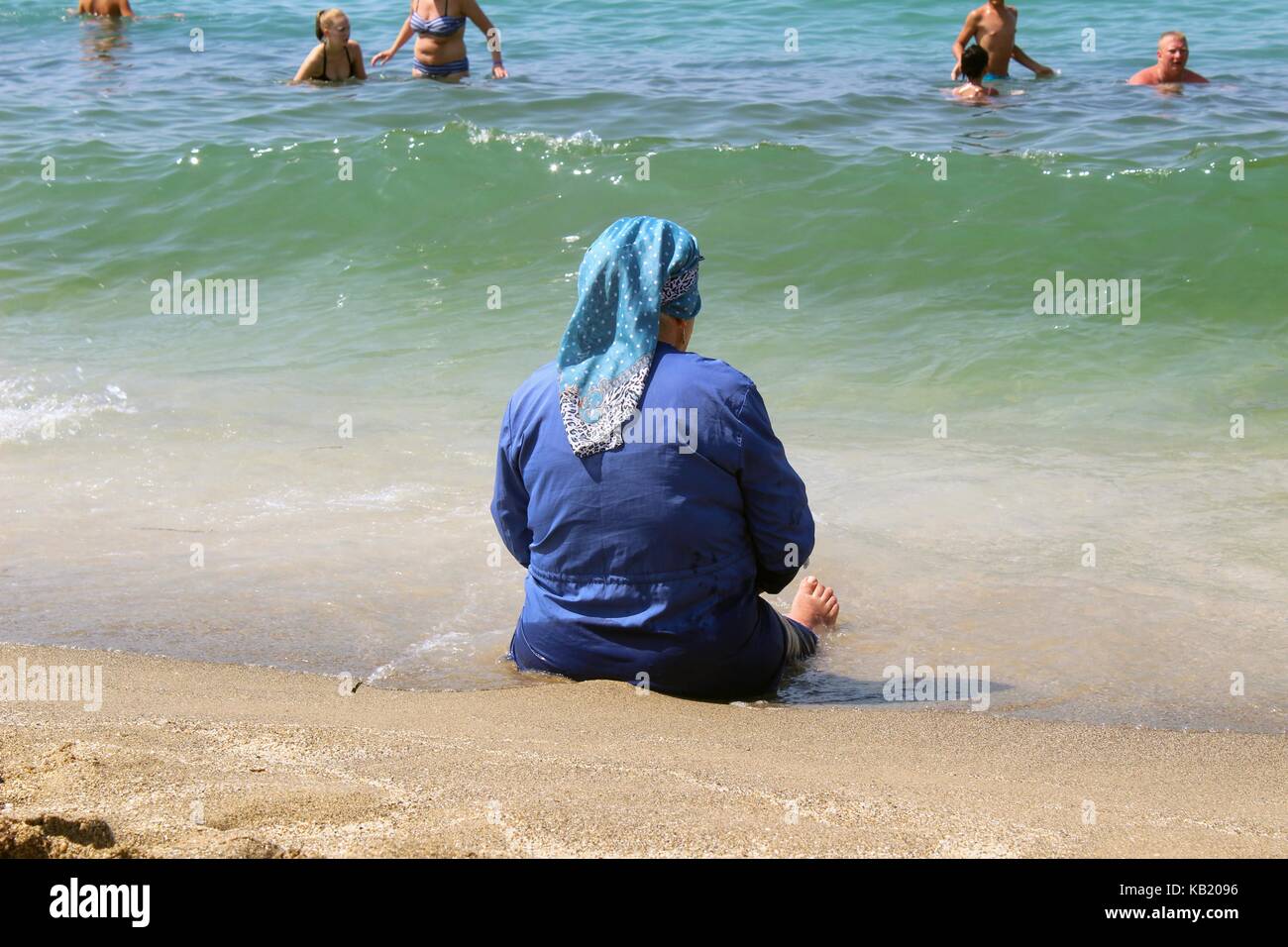 Juli, 2017 - Eine ältere muslimische Frau sitzt auf dem Sand in einem Surf Zone auf dem Hintergrund der Touristen aus Europa (Alanya, Türkei). Stockfoto