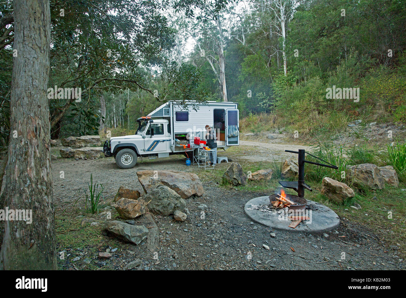 Camper außerhalb Kochen auf Gas Herd neben Allradantrieb Wohnmobil im Wald mit Lagerfeuer im Vordergrund in der australischen National Park Stockfoto