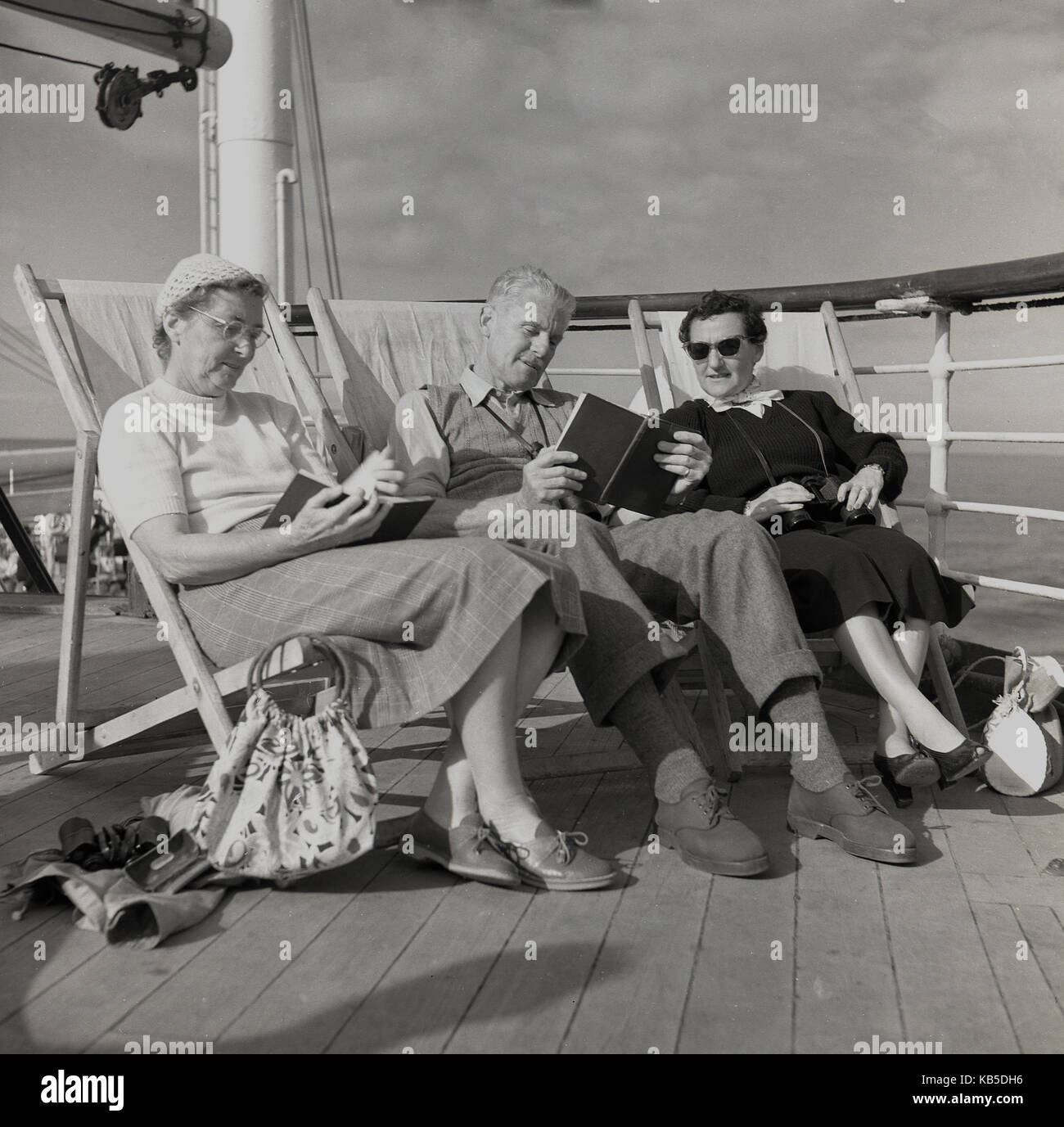 1950, historische, ältere Passagiere an Bord eines Schiffes Union-Castle sitzen in Liegestühlen auf dem Deck Bücher lesen und entspannen. Stockfoto