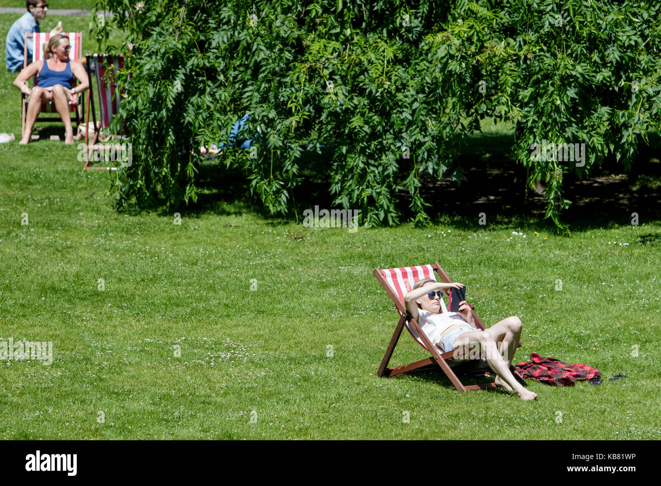 Eine Frau mit einem Buch und genießen Sie die warme Sonne und warmes Wetter wird dargestellt, sitzen in einem Liegestuhl in einem öffentlichen Park in Bath somerset England Vereinigtes Königreich Stockfoto