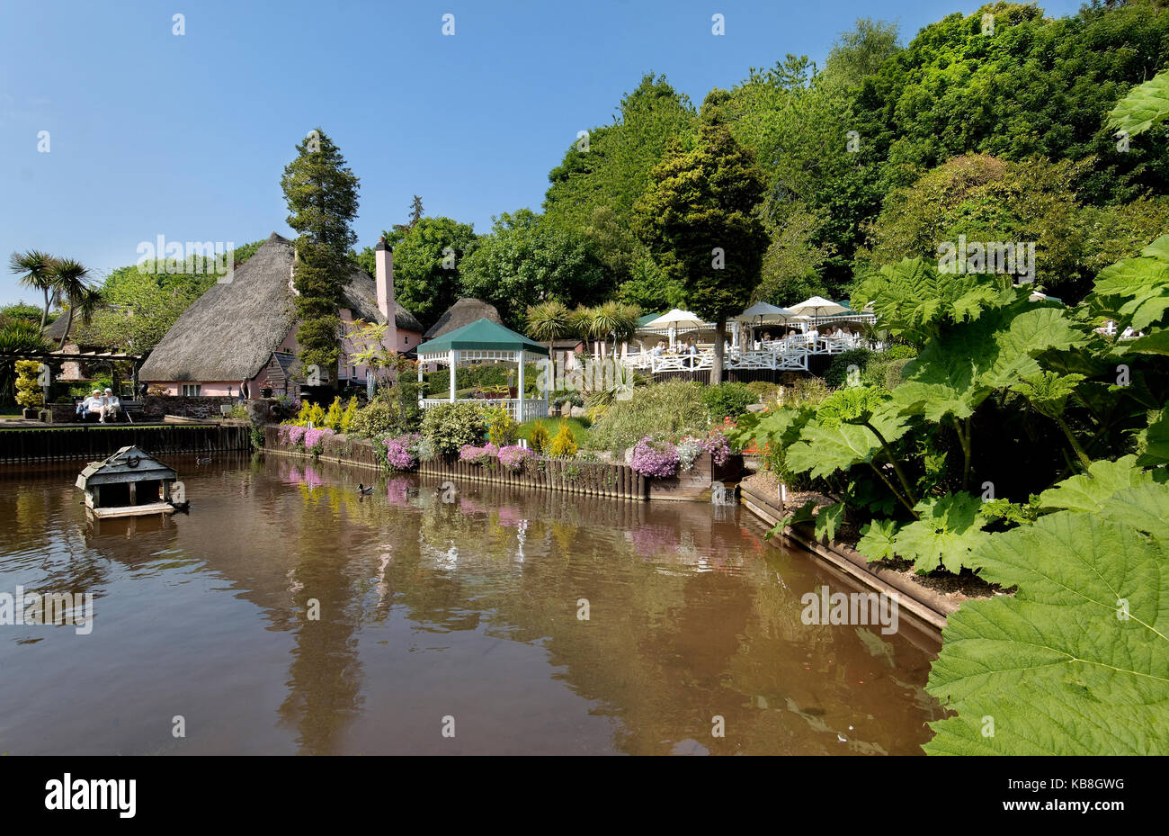 Rose Cottage Tea Gardens, cockington, Torquay Devon, Großbritannien - Blick über den Rasen und Stream mit, Kaffee Terrassen, strohgedeckten Kinderbett und Musikpavillon im Hintergrund Stockfoto