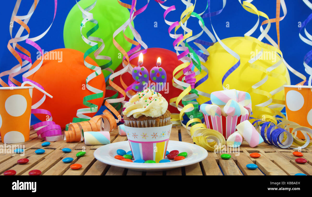 99 Geburtstag Kuchen mit Kerzen brennen auf der rustikalen Holztisch mit Hintergrund von bunten Luftballons plastik becher Bonbons auf der blauen Wand im Hintergrund Stockfoto