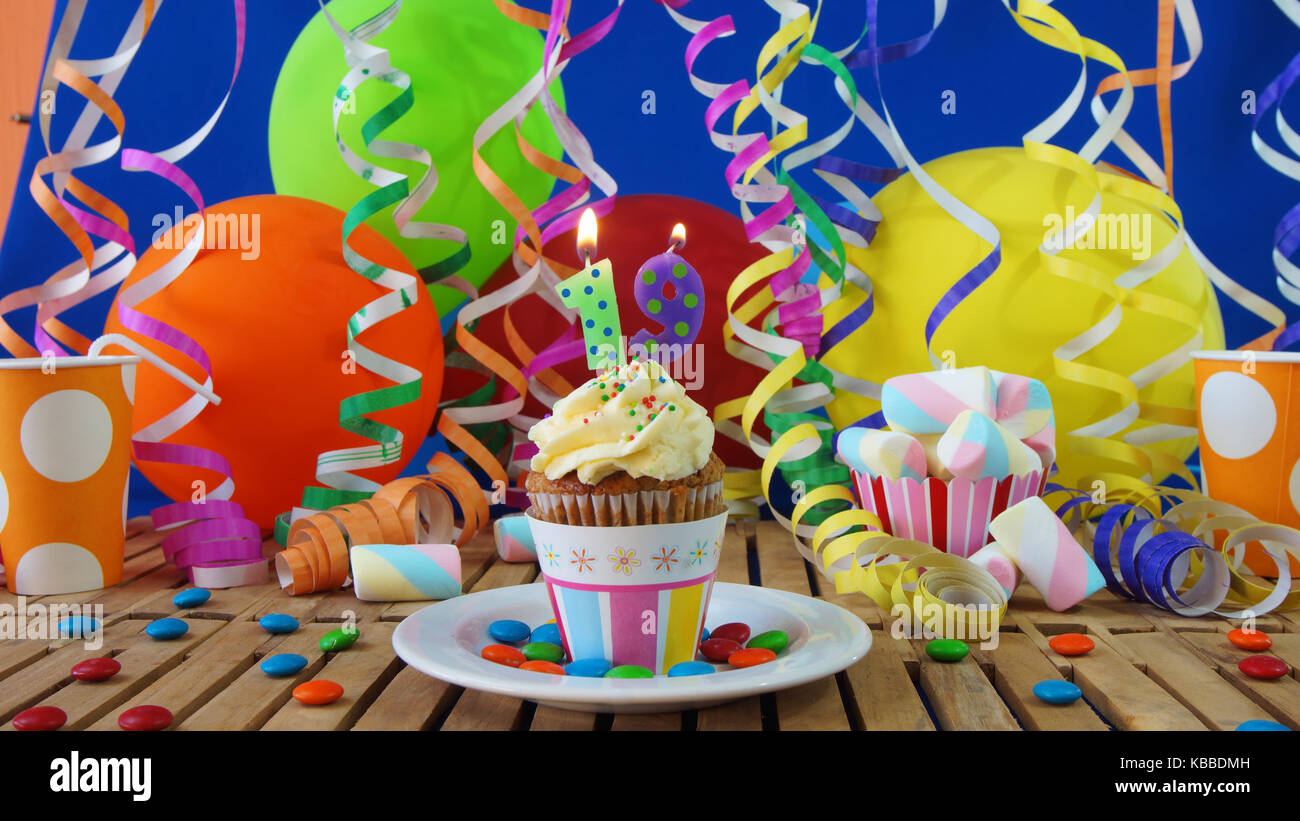 19 Geburtstag Kuchen mit Kerzen brennen auf der rustikalen Holztisch mit Hintergrund von bunten Luftballons plastik becher Bonbons auf der blauen Wand im Hintergrund Stockfoto