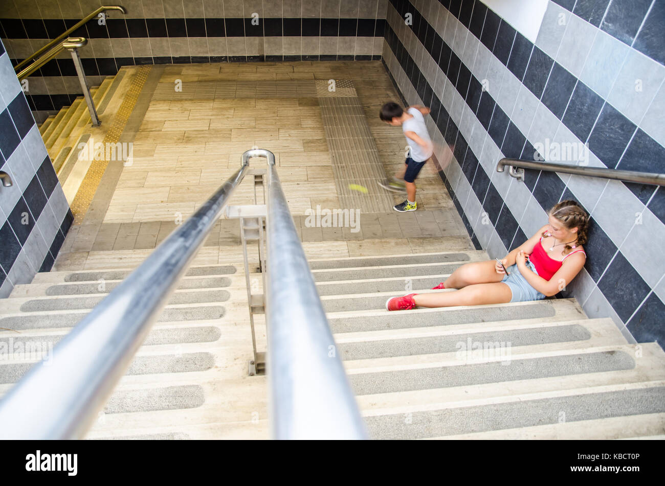 Warten auf die Bahn. Kaukasische Mädchen in Shorts und T-Shirts sitzen auf der Treppe von einem Bahnhof während der Beratung Ihr Smartphone. Kaukasische Junge ist Spielen Stockfoto