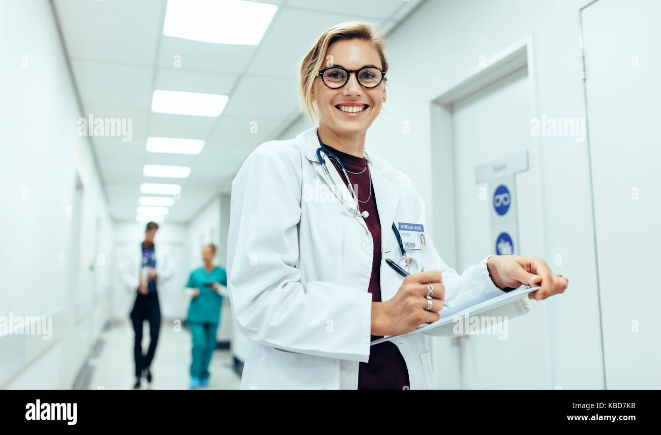 Portrait von glücklichen jungen weiblichen Arzt stehend im Flur mit Zwischenablage. Kaukasische Frau im Krankenhaus schreiben Rezept funktioniert. Stockfoto