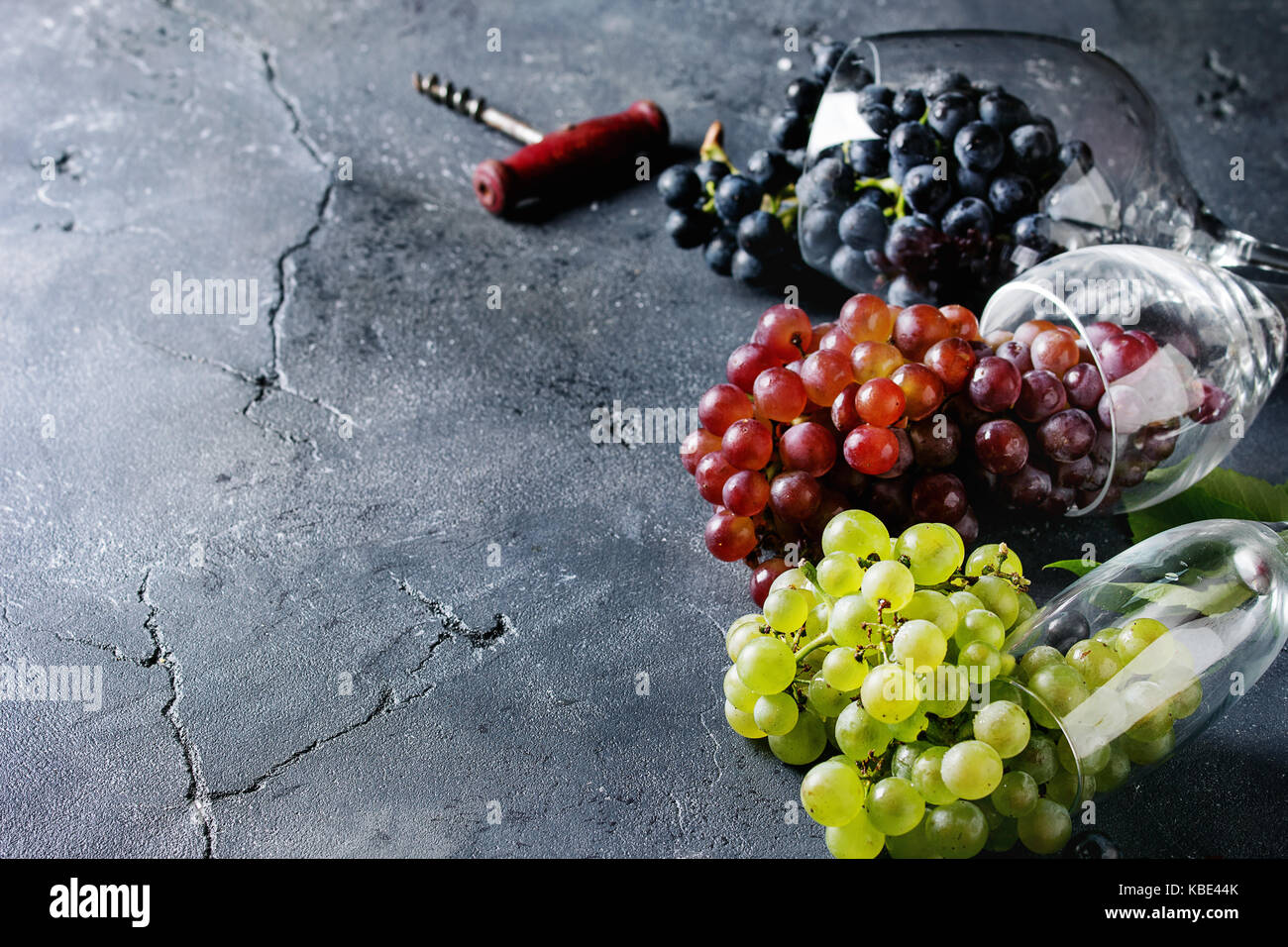 Vielzahl von drei Art frische reife Trauben dunkel Blau, Rot und Grün in verschiedenen Lügen Weingläser mit alten Korkenzieher über schwarzen Textur Hintergrund mit Stockfoto