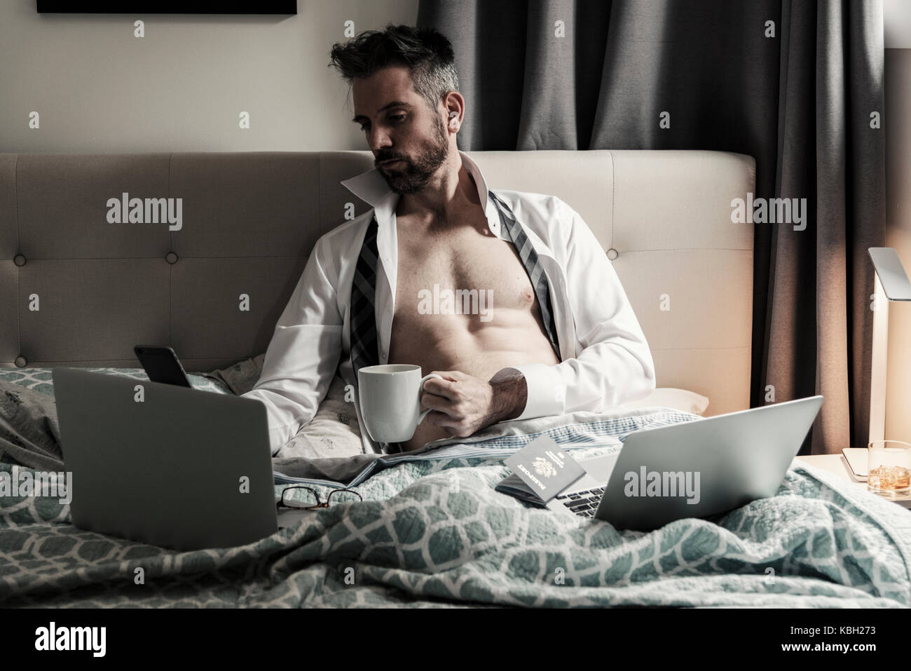 Gutaussehende kaukasische Männer, die im Bett mit mehreren Computern und mobilen Wireless-Geräten arbeiten, während sie eine Tasse Kaffee halten. Stockfoto